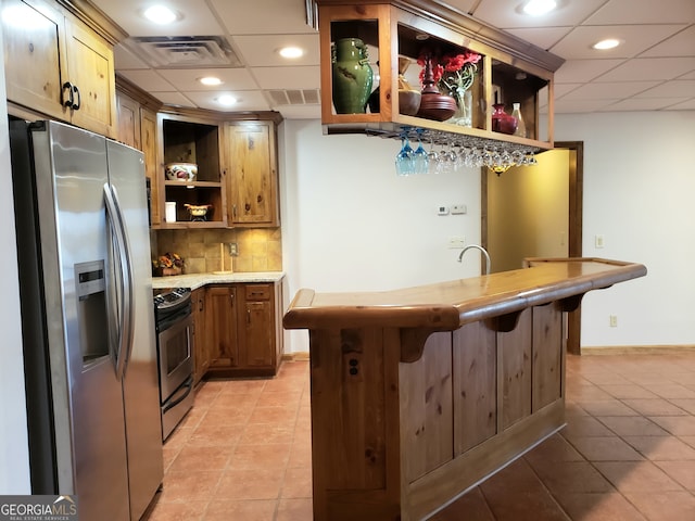 kitchen featuring backsplash, appliances with stainless steel finishes, a paneled ceiling, light tile floors, and a breakfast bar area