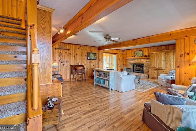 living room with light wood-type flooring, wooden walls, ceiling fan, beamed ceiling, and a stone fireplace