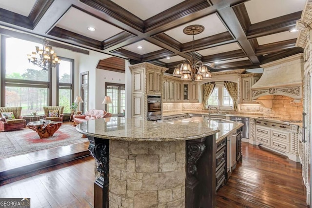 kitchen featuring coffered ceiling, an inviting chandelier, a kitchen island with sink, backsplash, and hanging light fixtures