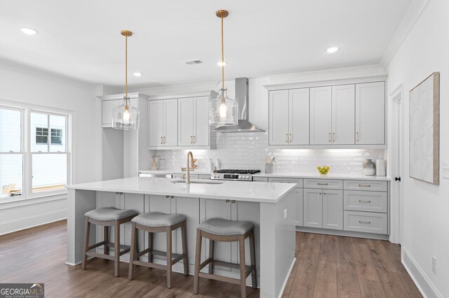 kitchen with a kitchen island with sink, wood finished floors, a sink, light countertops, and wall chimney range hood