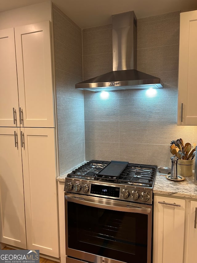 kitchen featuring stainless steel range with gas cooktop, white cabinets, light stone counters, and wall chimney range hood