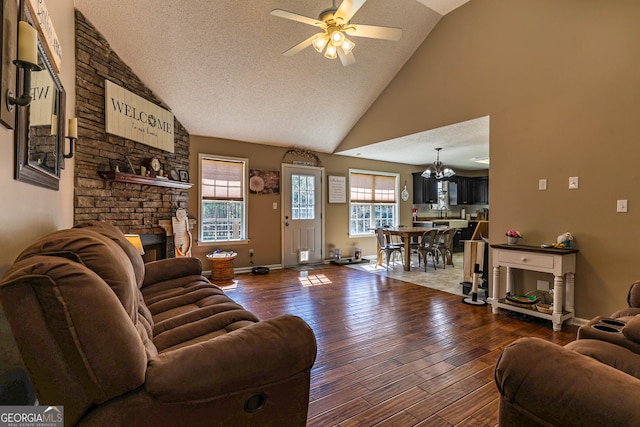 living room with dark wood-type flooring, a fireplace, a textured ceiling, high vaulted ceiling, and ceiling fan with notable chandelier