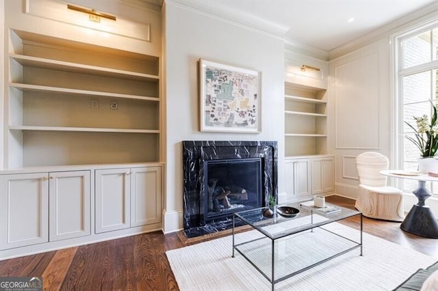 living room with built in features, dark wood-type flooring, ornamental molding, and a high end fireplace