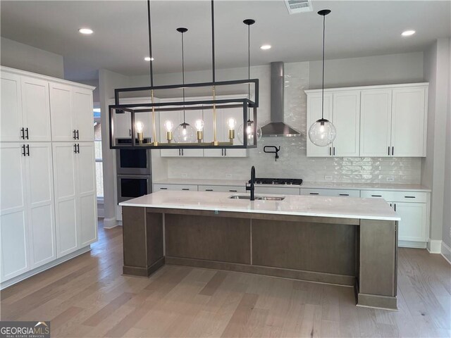 kitchen with double oven, backsplash, wall chimney range hood, decorative light fixtures, and a kitchen island with sink