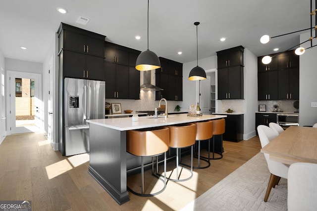 kitchen featuring sink, stainless steel fridge, hanging light fixtures, an island with sink, and wall chimney exhaust hood