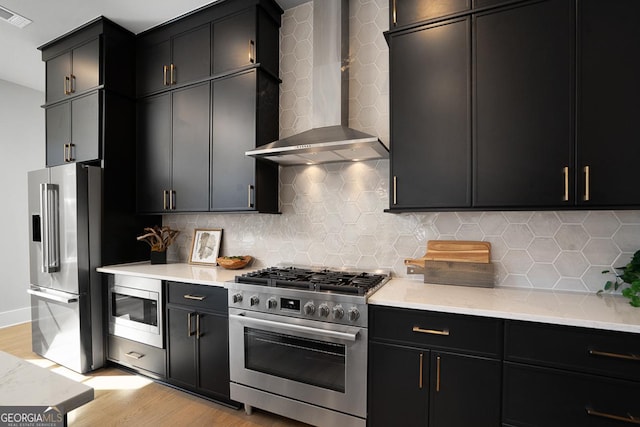 kitchen with tasteful backsplash, wall chimney range hood, premium appliances, and light hardwood / wood-style flooring