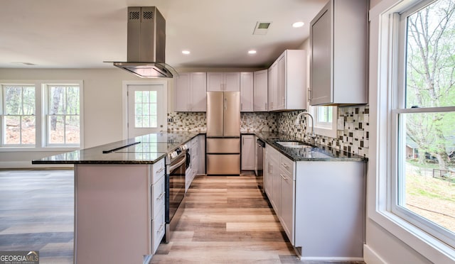 kitchen with appliances with stainless steel finishes, dark stone countertops, island exhaust hood, light hardwood / wood-style flooring, and sink