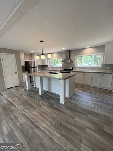 kitchen featuring appliances with stainless steel finishes, a kitchen island with sink, wall chimney exhaust hood, hardwood / wood-style floors, and pendant lighting