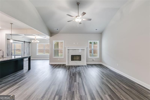 unfurnished living room featuring dark wood-type flooring, a high end fireplace, ceiling fan with notable chandelier, a barn door, and vaulted ceiling