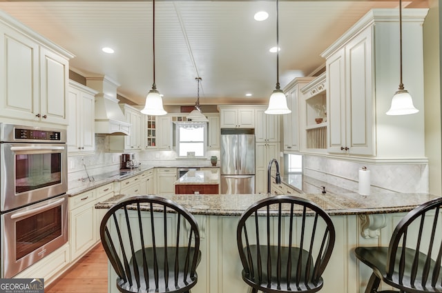 kitchen featuring appliances with stainless steel finishes, custom exhaust hood, backsplash, hanging light fixtures, and light hardwood / wood-style flooring