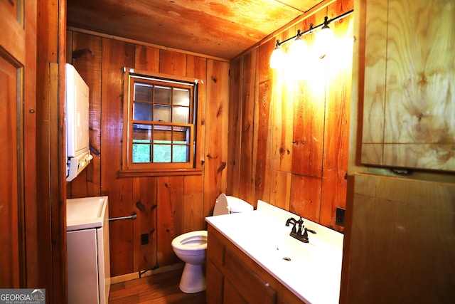 bathroom featuring wood walls, stacked washer / drying machine, wooden ceiling, vanity, and toilet