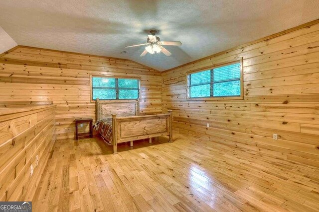 unfurnished bedroom featuring ceiling fan, light hardwood / wood-style floors, lofted ceiling, a textured ceiling, and wooden walls