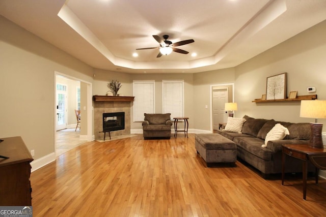 living room featuring ceiling fan, a tray ceiling, a tile fireplace, and light hardwood / wood-style flooring