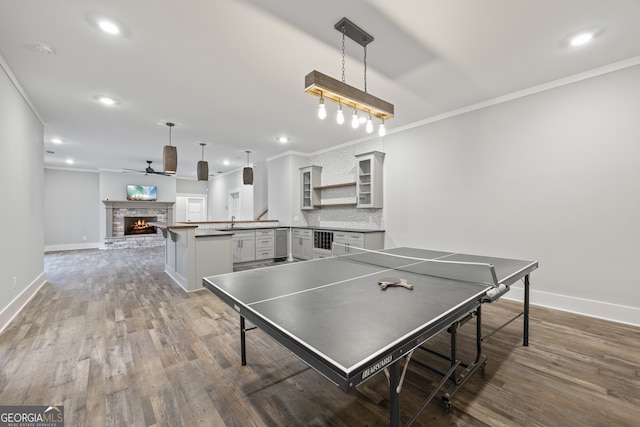 recreation room featuring ornamental molding, wood-type flooring, a stone fireplace, and ceiling fan