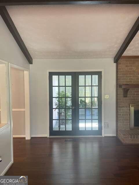 unfurnished living room with french doors, vaulted ceiling with beams, a brick fireplace, dark hardwood / wood-style flooring, and brick wall