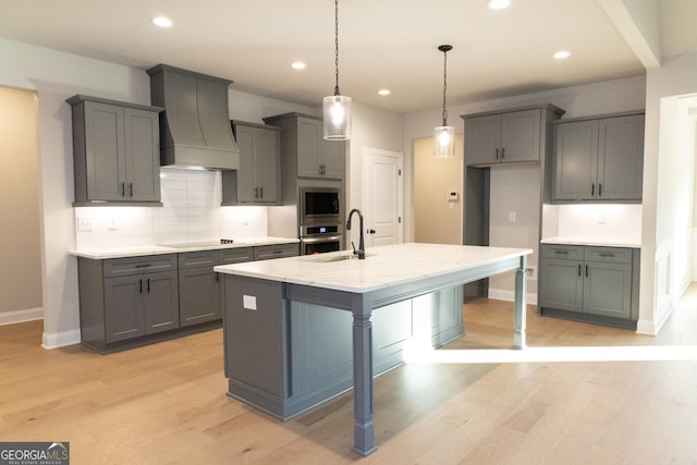 kitchen featuring stainless steel appliances, gray cabinets, custom range hood, a kitchen island with sink, and a sink