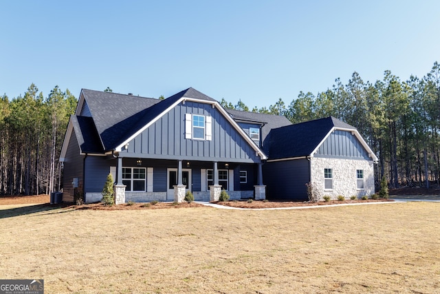 view of front of house with stone siding, roof with shingles, covered porch, a front lawn, and board and batten siding