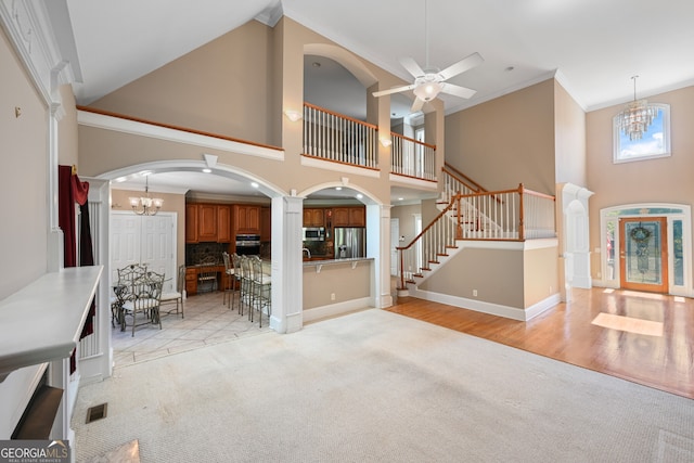 unfurnished living room featuring ceiling fan with notable chandelier, light carpet, crown molding, high vaulted ceiling, and ornate columns