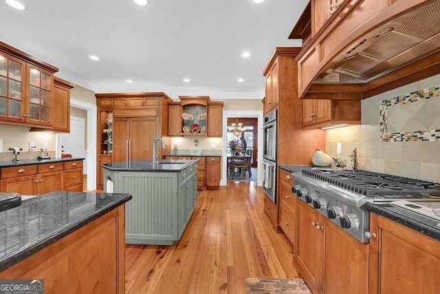 kitchen with a center island with sink, custom range hood, stainless steel appliances, sink, and light wood-type flooring