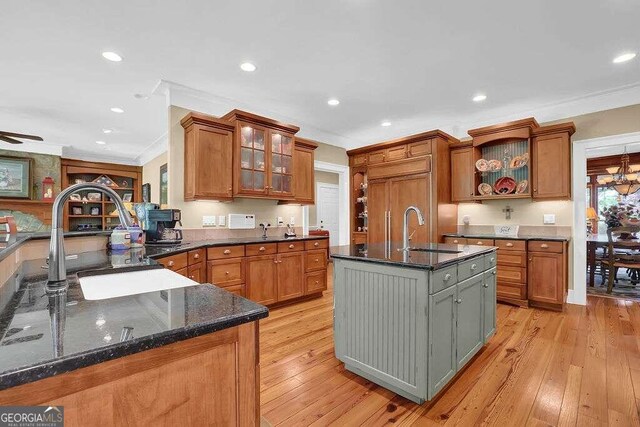 kitchen featuring light wood-type flooring, dark stone counters, ceiling fan with notable chandelier, an island with sink, and sink