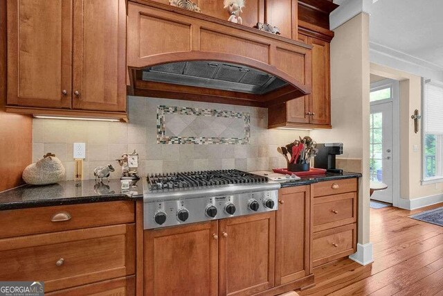 kitchen featuring stainless steel gas stovetop, dark stone countertops, light hardwood / wood-style flooring, premium range hood, and tasteful backsplash