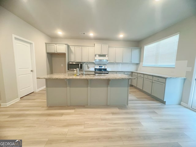 kitchen featuring light stone counters, an island with sink, light wood-type flooring, and appliances with stainless steel finishes