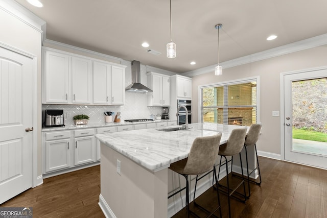 kitchen featuring white cabinets, an island with sink, hanging light fixtures, and wall chimney range hood