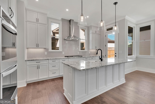 kitchen with pendant lighting, wall chimney range hood, a kitchen island with sink, white cabinetry, and dark hardwood / wood-style flooring