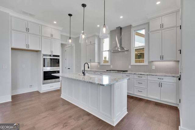 kitchen with white cabinets, an island with sink, hanging light fixtures, and wall chimney range hood