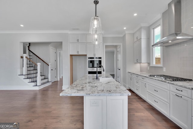 kitchen with pendant lighting, wall chimney range hood, stainless steel appliances, an island with sink, and white cabinets