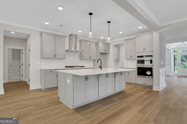 kitchen featuring double oven, a kitchen island with sink, light wood-type flooring, and wall chimney range hood