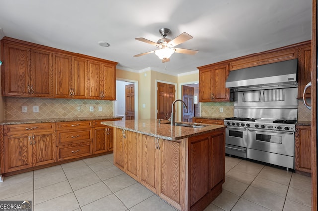 kitchen featuring wall chimney exhaust hood, decorative backsplash, double oven range, crown molding, and a center island with sink