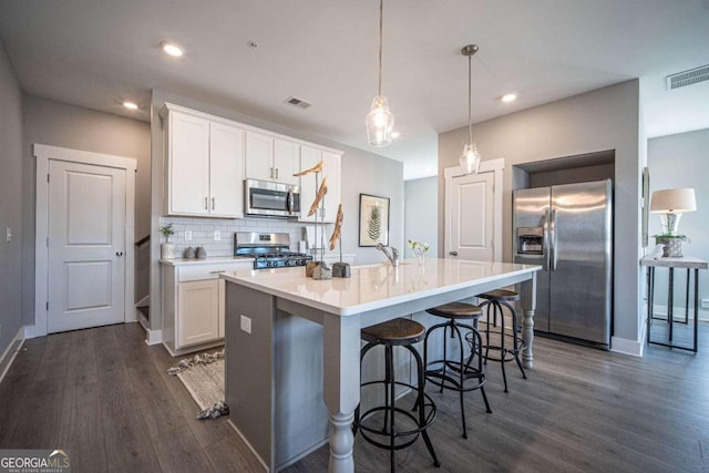 kitchen with white cabinetry, hanging light fixtures, appliances with stainless steel finishes, dark hardwood / wood-style flooring, and a kitchen island with sink