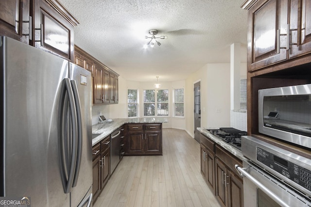 kitchen with appliances with stainless steel finishes, light stone counters, dark brown cabinetry, pendant lighting, and light hardwood / wood-style floors