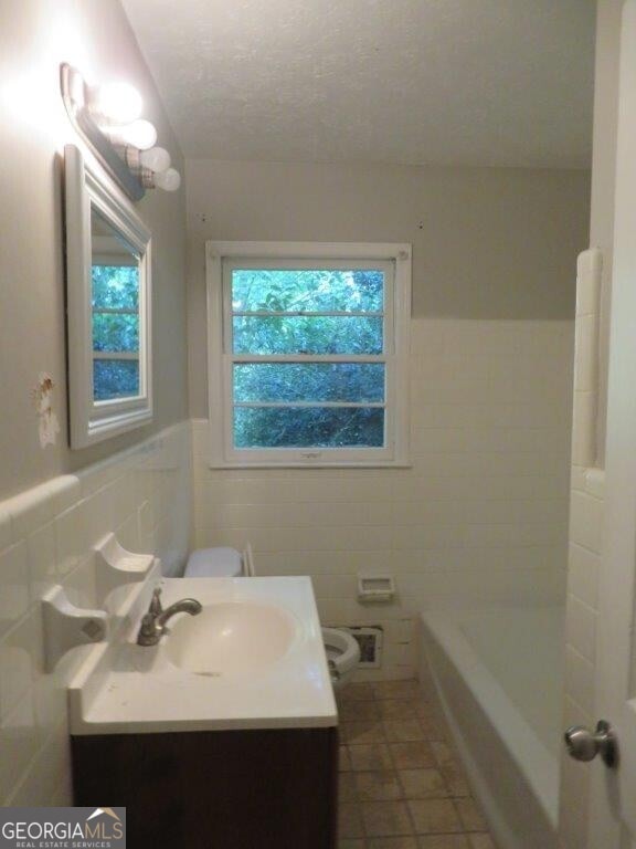 bathroom with a tub to relax in, a textured ceiling, vanity, tile walls, and toilet