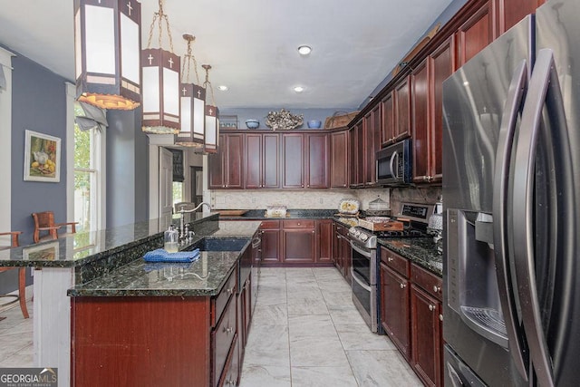 kitchen with backsplash, dark stone counters, hanging light fixtures, an island with sink, and appliances with stainless steel finishes