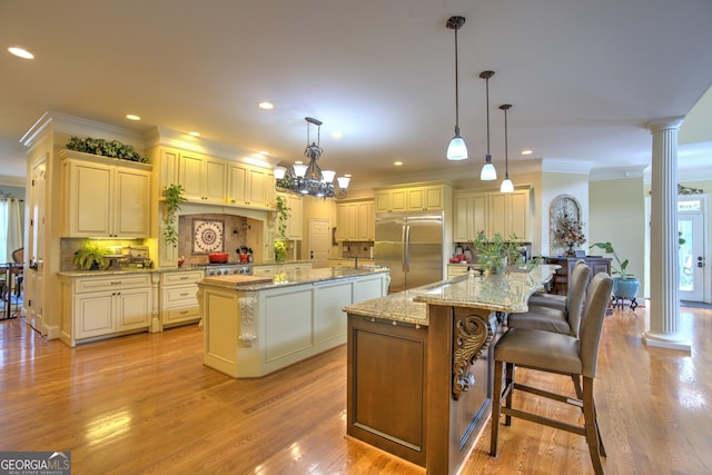 kitchen with built in fridge, hanging light fixtures, a center island with sink, and ornate columns