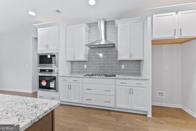 kitchen with decorative backsplash, appliances with stainless steel finishes, wall chimney exhaust hood, light hardwood / wood-style flooring, and white cabinetry