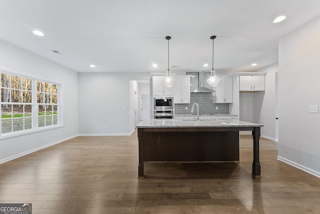 kitchen featuring white cabinetry, wall chimney exhaust hood, light stone counters, a kitchen island with sink, and appliances with stainless steel finishes