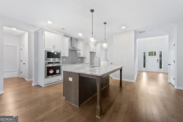 kitchen with sink, stainless steel appliances, wall chimney range hood, a center island with sink, and white cabinets