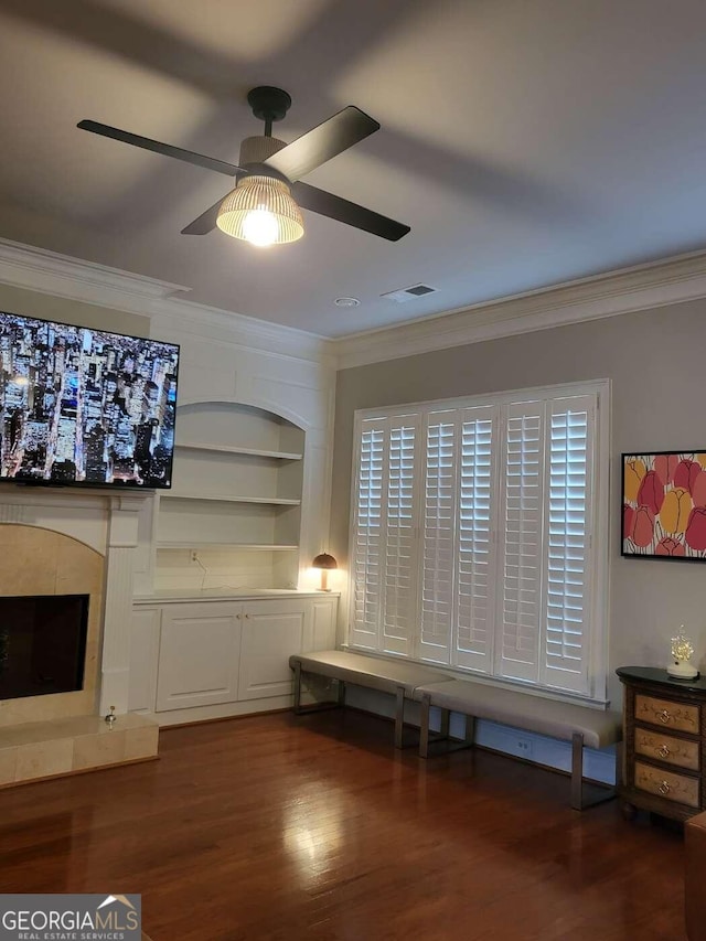 unfurnished room featuring ornamental molding, a tiled fireplace, built in shelves, wood-type flooring, and ceiling fan