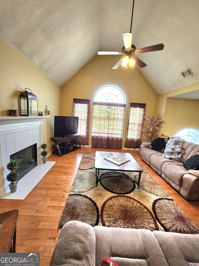 living room with a tiled fireplace, vaulted ceiling, light wood-type flooring, a textured ceiling, and ceiling fan