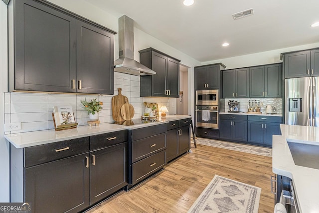 kitchen featuring tasteful backsplash, wall chimney range hood, stainless steel appliances, light stone countertops, and light hardwood / wood-style flooring