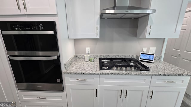 kitchen with dobule oven black, stainless steel gas cooktop, wall chimney exhaust hood, light stone counters, and white cabinetry