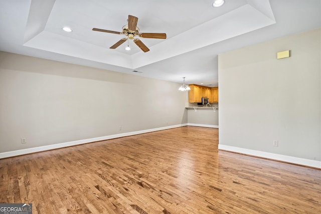 unfurnished living room featuring ceiling fan with notable chandelier, a raised ceiling, and light wood-type flooring