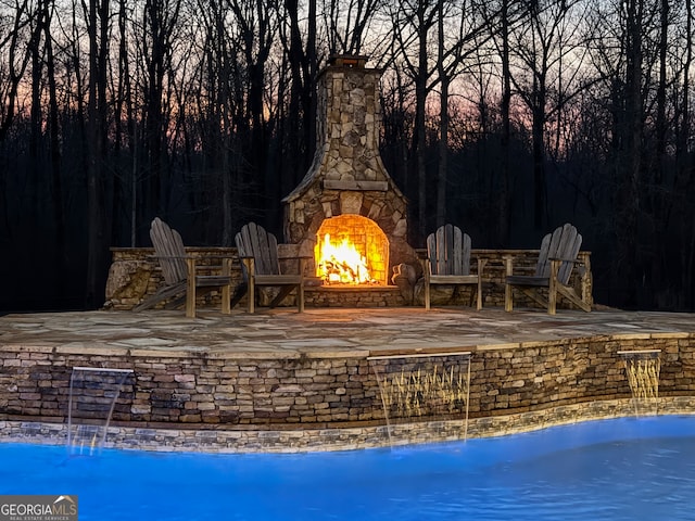 pool at dusk with an outdoor stone fireplace and a patio area