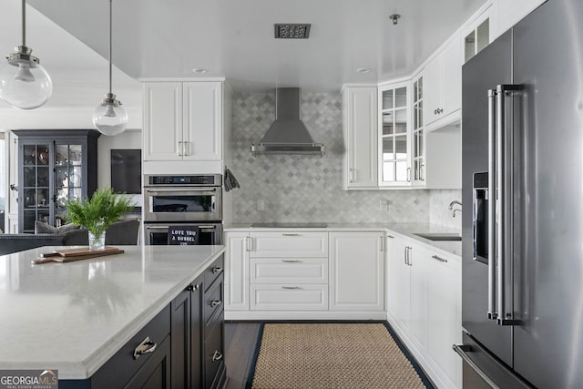 kitchen with light countertops, appliances with stainless steel finishes, glass insert cabinets, a sink, and wall chimney range hood