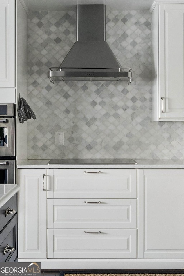 kitchen featuring tasteful backsplash, light countertops, wall chimney range hood, double oven, and white cabinetry