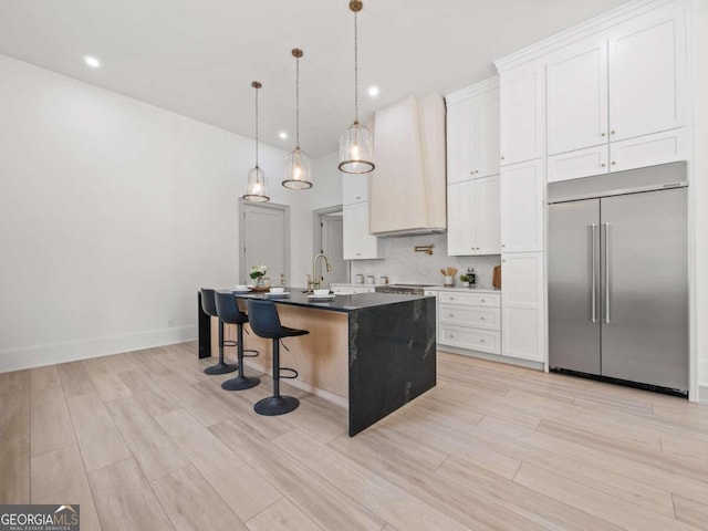 kitchen featuring built in fridge, a center island with sink, white cabinets, and hanging light fixtures