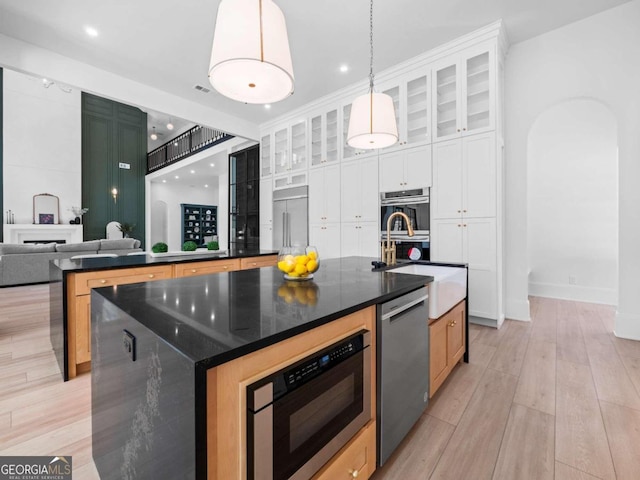 kitchen featuring stainless steel appliances, decorative light fixtures, light hardwood / wood-style flooring, white cabinetry, and an island with sink